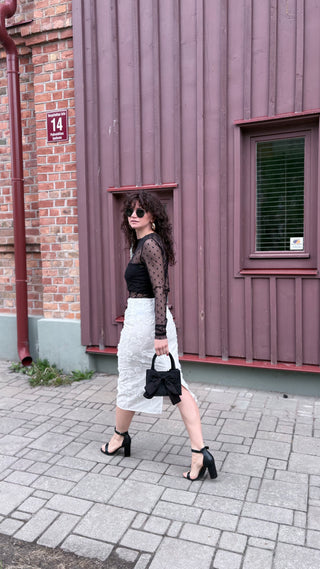 A woman walking outdoors on a paved street in front of a purple wooden building, wearing a black sheer long-sleeve top, a white textured midi skirt, black high-heeled sandals, and carrying a small black handbag.