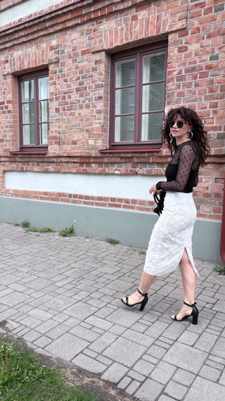 A woman wearing a black sheer long-sleeve top and a white lace midi skirt with a side slit, walking on a paved sidewalk in front of a brick building.
