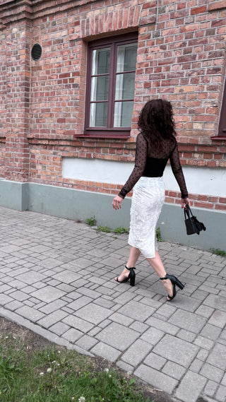 A woman walking on a paved sidewalk in front of a red brick building, wearing a black sheer long-sleeve top, a white textured midi skirt, and black high heels, while carrying a black handbag.