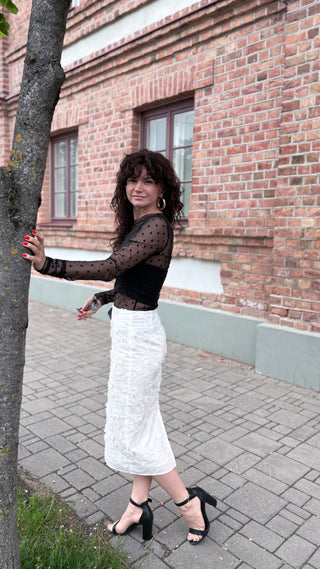 A woman posing outdoors in front of a brick building, wearing a black sheer long-sleeve top with polka dots and a white textured midi skirt, paired with black high-heeled sandals.
