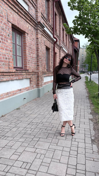A woman stands on a paved sidewalk next to a brick building, wearing a black sheer long-sleeve top with a black corset-style midriff and a white textured midi skirt, holding a black handbag.