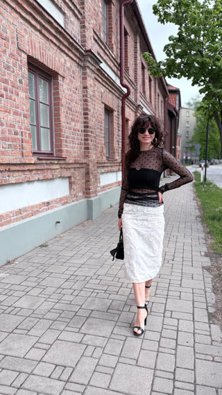 A woman stands on a paved sidewalk in front of a brick building, wearing a sheer black long-sleeve top with a black bandeau and a white textured midi skirt, holding a black handbag.