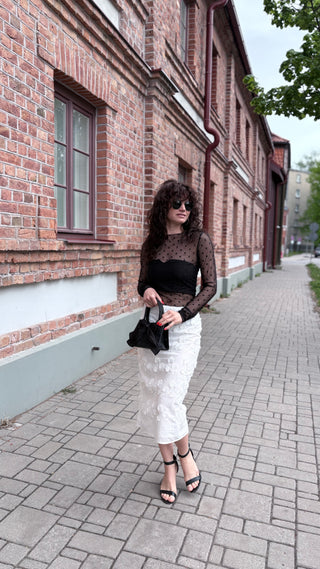 A woman stands on a paved sidewalk next to a brick building, wearing a black sheer long-sleeve top, a white textured midi skirt, black strappy heels, and holding a black handbag.