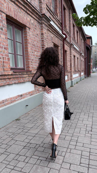 A woman is photographed from behind walking down a paved street next to a brick building. She is wearing a sheer black long-sleeve top over a black bandeau, a white textured midi skirt with a front slit, and black ankle boots, while carrying a black handbag.