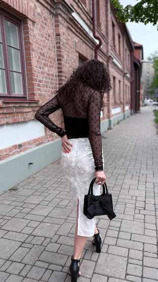 A woman stands on a paved street in front of a brick building, wearing a sheer black long-sleeve top with black polka dots, a black cropped bodysuit, a white lace midi skirt with a side slit, and holding a black handbag.