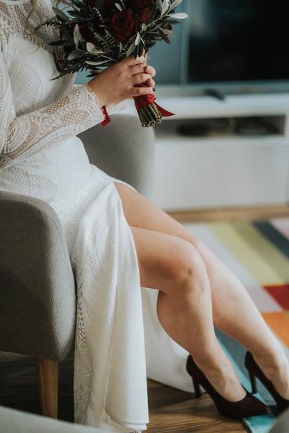 lady with a beautiful bouquet of flowers sitting on a sofa in her bordeaux shoes and lace wedding dress